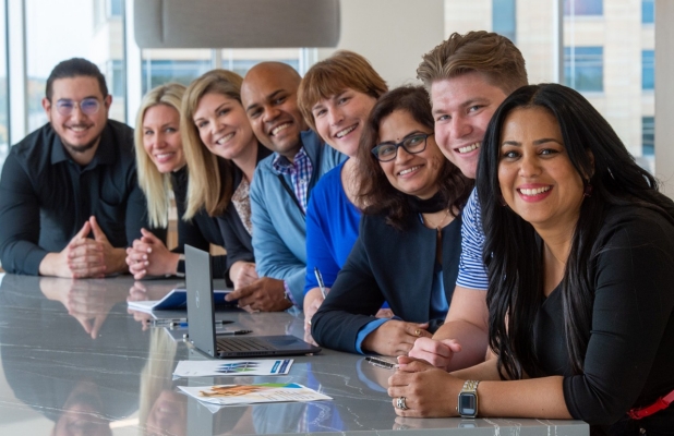 Eight people leaning on a table and smilinng towards you. 